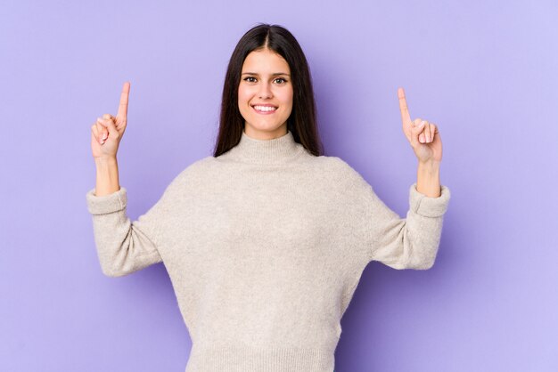 Young caucasian woman isolated on purple wall indicates with both fore fingers up showing a blank space.