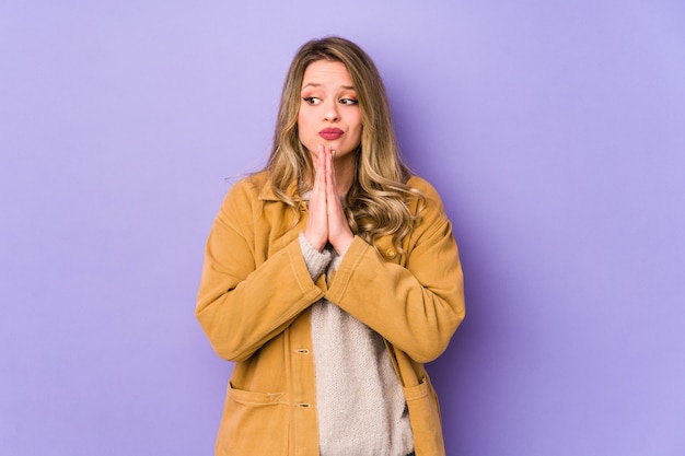 Young caucasian woman isolated on purple wall praying, showing devotion, religious person looking for divine inspiration.