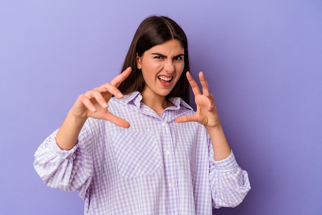 Young caucasian woman isolated on purple wall showing claws imitating a cat, aggressive gesture
