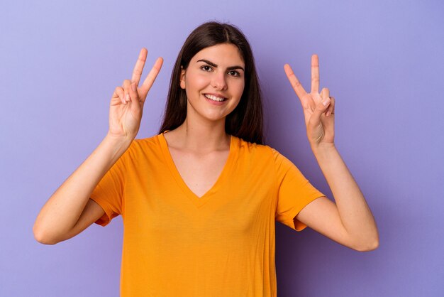 Young caucasian woman isolated on purple wall showing victory sign and smiling broadly.