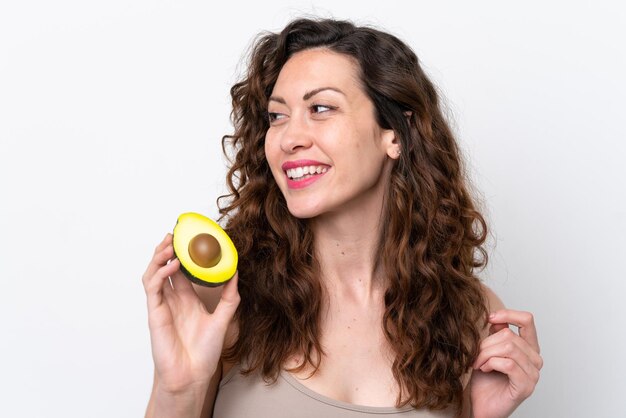 Young caucasian woman isolated on white background holding an avocado while smiling Close up portrait