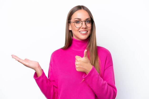 Young caucasian woman isolated on white background holding copyspace imaginary on the palm to insert an ad and with thumbs up