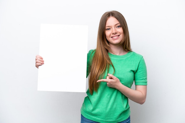 Giovane donna caucasica isolata su sfondo bianco in possesso di un cartello vuoto con felice espressione e indicandolo