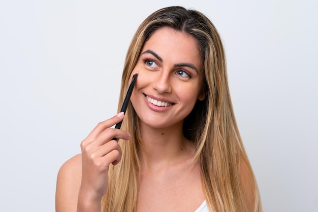 Young caucasian woman isolated on white background holding makeup brush and whit happy expression