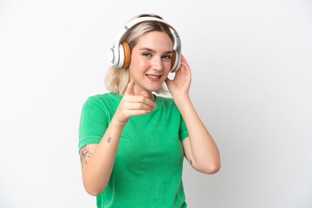 Foto giovane donna caucasica isolata su sfondo bianco ascoltando musica e indicando la parte anteriore