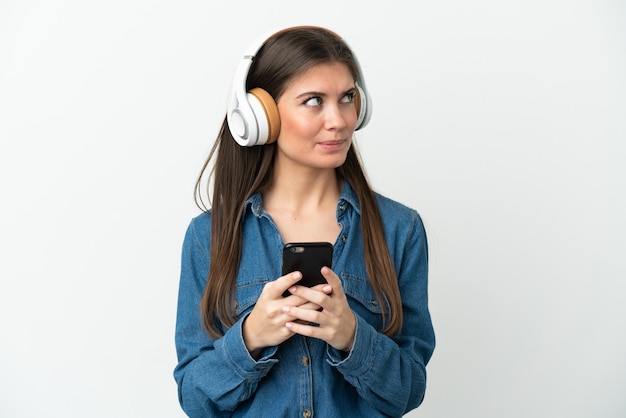 Young caucasian woman isolated on white background listening music with a mobile and thinking