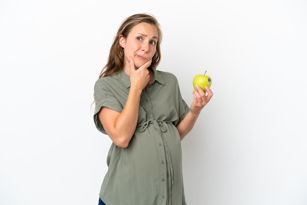 Young caucasian woman isolated on white background pregnant and holding an apple while thinking