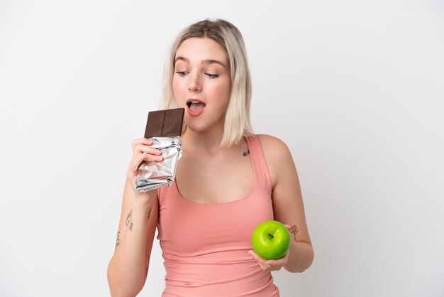 Young caucasian woman isolated on white background taking a chocolate tablet in one hand and an apple in the other