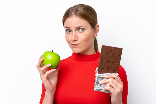 Молодая кавказская женщина изолирована на белом фоне, принимая шоколадную таблетку в одной руке и яблоко в другой