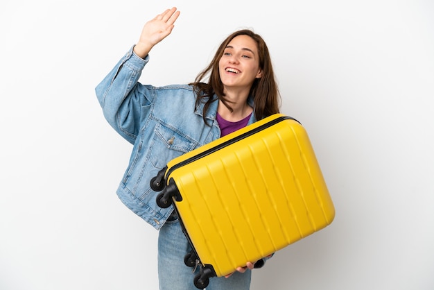 Young caucasian woman isolated on white background in vacation with travel suitcase and saluting