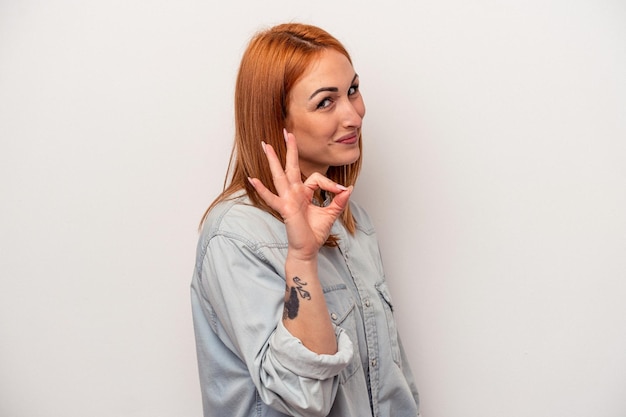 Young caucasian woman isolated on white background winks an eye and holds an okay gesture with hand