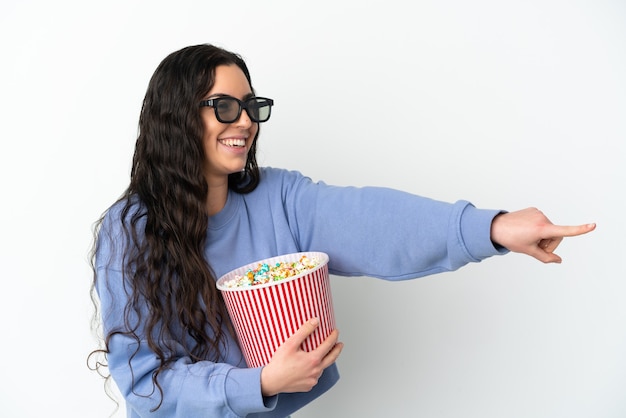 Young caucasian woman isolated on white background with 3d glasses and holding a big bucket of popcorns while pointing away