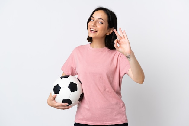 Giovane donna caucasica isolata su sfondo bianco con pallone da calcio e facendo segno ok