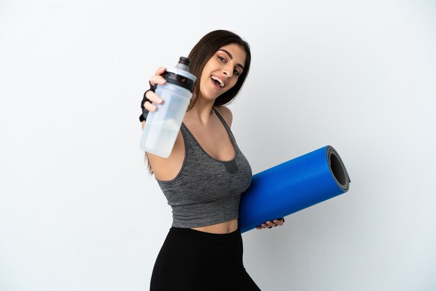 Молодая кавказская женщина изолирована на белом фоне со спортивной бутылкой с водой и с циновкой