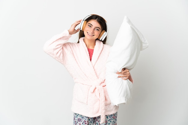 Young caucasian woman isolated on white wall in pajamas and holding a pillow and listening music