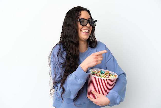 Young caucasian woman isolated on white wall with 3d glasses and holding a big bucket of popcorns while pointing front