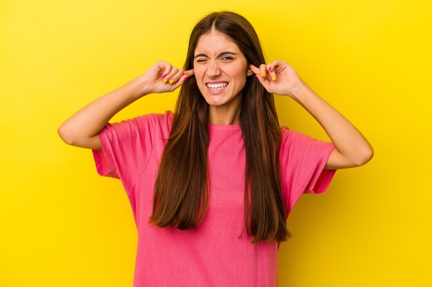 Young caucasian woman isolated on yellow background covering ears with fingers, stressed and desperate by a loudly ambient.