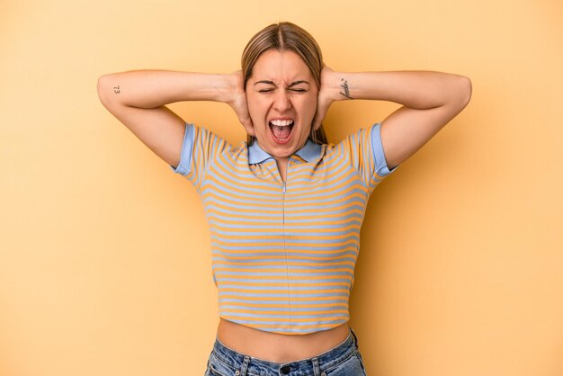 Young caucasian woman isolated on yellow background covering ears with hands trying not to hear too loud sound.