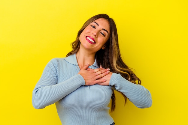 Young caucasian woman isolated on yellow background has friendly expression, pressing palm to chest. Love concept.