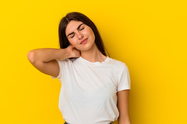 Giovane donna caucasica isolata su sfondo giallo che ha un dolore al collo a causa dello stress, massaggiandolo e toccandolo con la mano.