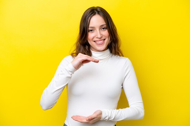 Young caucasian woman isolated on yellow background holding copyspace imaginary on the palm to insert an ad