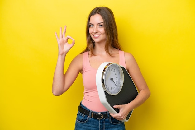 Young caucasian woman isolated on yellow background holding a weighing machine and doing OK sign