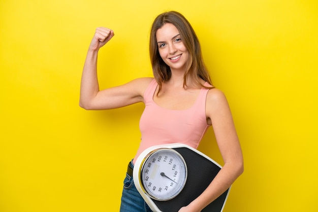 Young caucasian woman isolated on yellow background holding a weighing machine and doing strong gesture