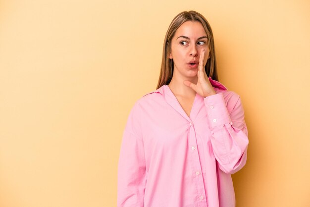 Young caucasian woman isolated on yellow background is saying a secret hot braking news and looking aside