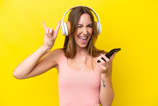 Young caucasian woman isolated on yellow background listening music with a mobile making rock gesture