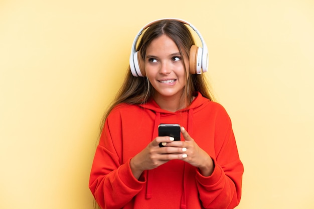 携帯電話で音楽を聴いて、思考と黄色の背景に分離された若い白人女性