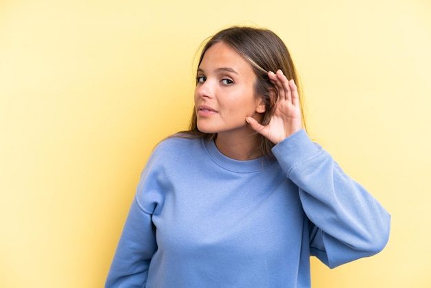 Photo young caucasian woman isolated on yellow background listening to something by putting hand on the ear