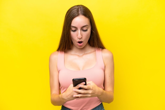 Молодая кавказская женщина, изолированная на желтом фоне, смотрит в камеру, используя мобильный телефон, с удивленным выражением лица