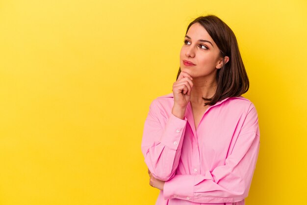 Giovane donna caucasica isolata su sfondo giallo guardando lateralmente con espressione dubbiosa e scettica.