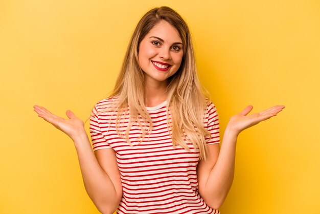 Young caucasian woman isolated on yellow background makes scale with arms feels happy and confident