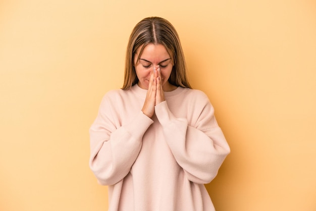 Young caucasian woman isolated on yellow background praying, showing devotion, religious person looking for divine inspiration.