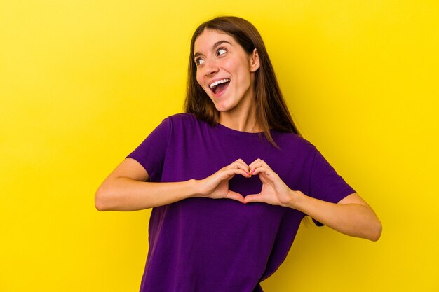 Young caucasian woman isolated on yellow background smiling and showing a heart shape with hands