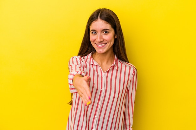 Young caucasian woman isolated on yellow background stretching hand at camera in greeting gesture.