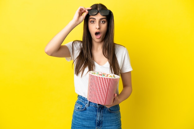 Young caucasian woman isolated on yellow background surprised with 3d glasses and holding a big bucket of popcorns