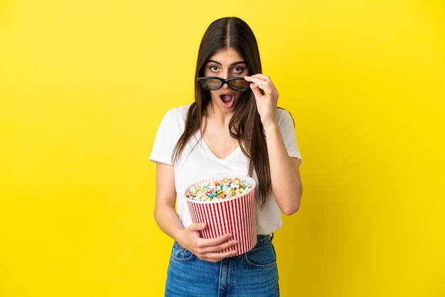 Young caucasian woman isolated on yellow background surprised with 3d glasses and holding a big bucket of popcorns