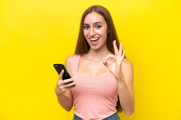 Young caucasian woman isolated on yellow background using mobile phone and doing ok sign