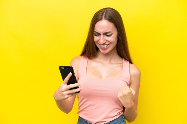 Young caucasian woman isolated on yellow background using mobile phone and doing victory gesture