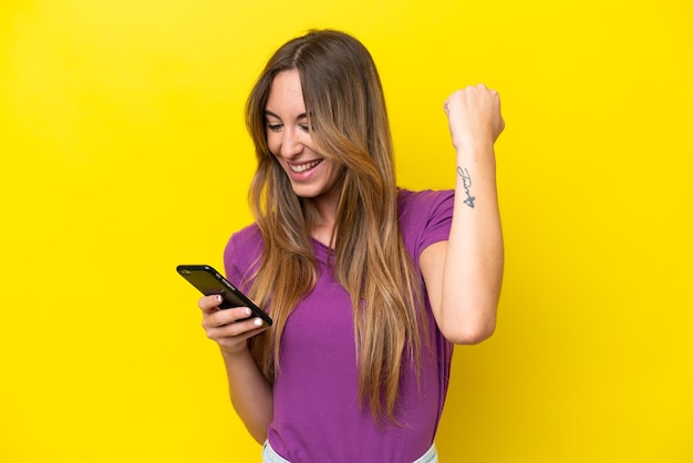 Young caucasian woman isolated on yellow background using mobile phone and doing victory gesture