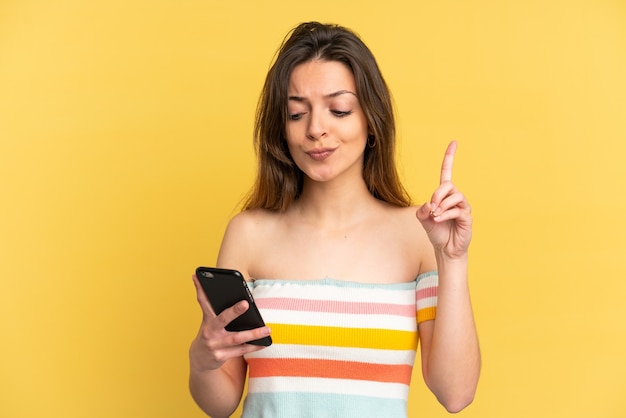 Young caucasian woman isolated on yellow background using mobile phone and lifting finger