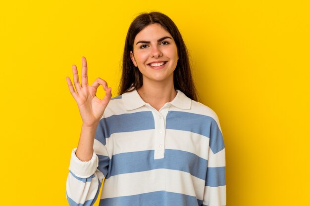 Молодая кавказская женщина, изолированная на желтом фоне, подмигивает и держит рукой хороший жест.