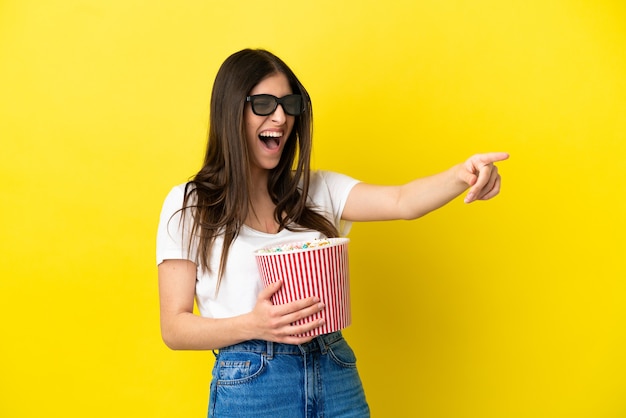 Young caucasian woman isolated on yellow background with 3d glasses and holding a big bucket of popcorns while pointing away