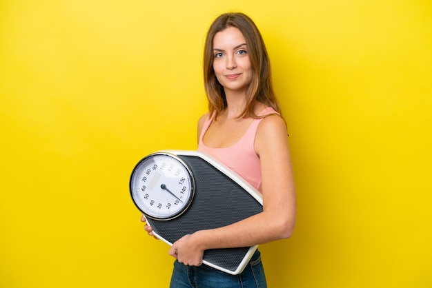 Young caucasian woman isolated on yellow background with weighing machine