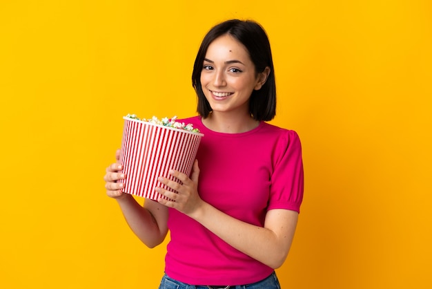 Giovane donna caucasica isolata sulla parete gialla che tiene un grande secchio di popcorn