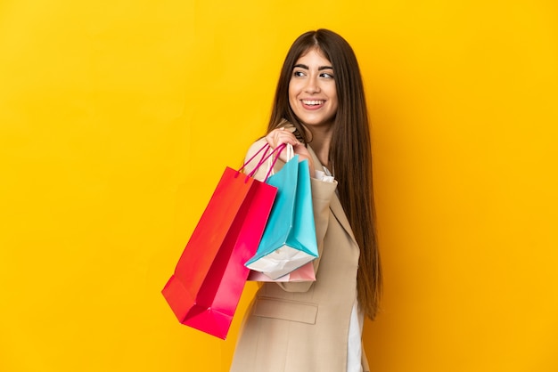 Young caucasian woman isolated on yellow wall holding shopping bags and looking back