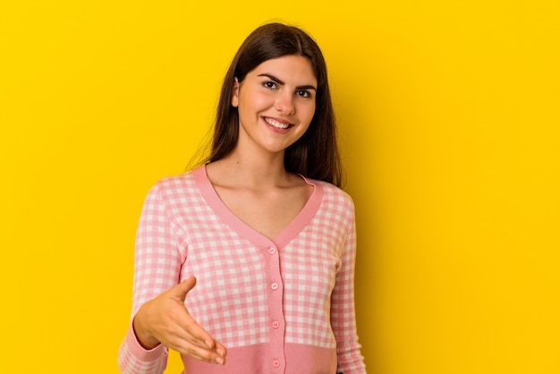 Young caucasian woman isolated on yellow wall stretching hand at camera in greeting gesture