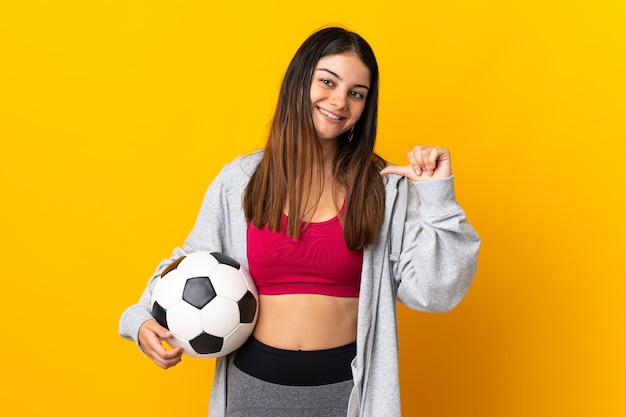 Молодая кавказская женщина изолирована на желтом с футбольным мячом и гордится собой